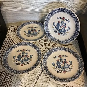 May include: A set of four vintage ceramic plates with a blue and white floral design. Each plate features a central image of two figures in traditional clothing. The plates are arranged on a white lace tablecloth.