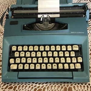 Może przedstawiać: Niebieska maszynka do pisania Smith-Corona z włożonym białym rolką papieru. Maszynka do pisania ma standardową klawiaturę QWERTY z czarnymi klawiszami i kremowymi napisami.