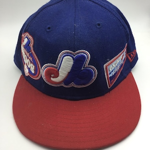 Peut inclure: Une casquette de baseball bleue et rouge avec plusieurs patchs, dont un grand logo "J", un logo "Los Angeles" plus petit et un logo "New Era".