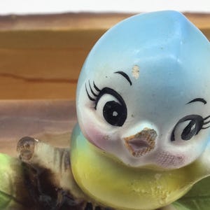 May include: A ceramic bird planter with a blue, yellow, and white painted design. The bird has large eyes and a small beak. The planter is sitting on a brown branch.