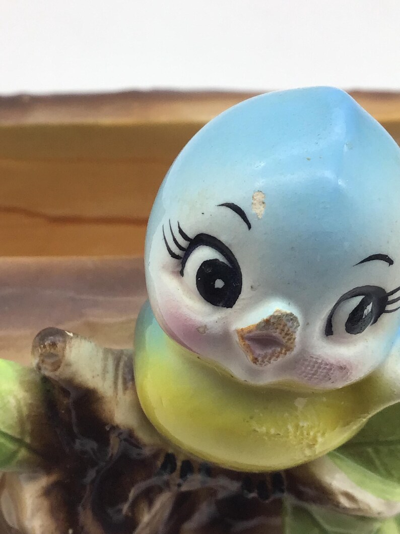 May include: A ceramic bird planter with a blue, yellow, and white painted design. The bird has large eyes and a small beak. The planter is sitting on a brown branch.