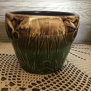 Op de afbeelding: Een keramische plantenbak met een bruine rand en een tweekleurige bruin en groene buitenkant. De plantenbak heeft een textuurontwerp met bloemen en geometrische patronen. De plantenbak staat op een crèmekleurig gehaakt kleedje.