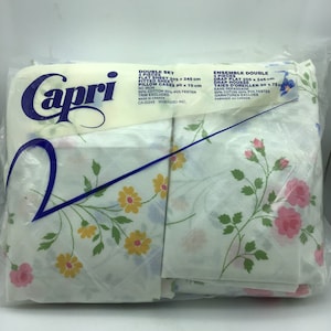 May include: A Capri brand double bed sheet set with a floral pattern in shades of pink, yellow, and blue. The set includes a flat sheet, fitted sheet, and two pillowcases. The packaging includes the text "Double Set 4 Pieces Flat Sheet 205 x 245 cm Fitted Sheet 205 x 245 cm Pillow Cases 60 x 75 cm No Iron 50% Cotton 50% Polyester Trim Excluded Made in Canada CA-00245 WABASSO INC."