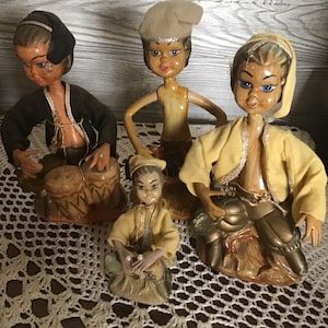Op de afbeelding: Vier vintage beeldjes van mensen in traditionele kledij. De beeldjes zijn beschilderd met gedetailleerde gelaatstrekken en dragen hoeden en jassen. Eén figuur speelt drums, een andere houdt een klein voorwerp vast.