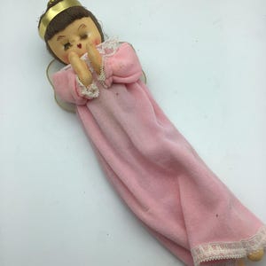 Könnte beinhalten: Eine Vintage-Engelfigur mit einem rosa Kleid, weißen Spitzenbesatz und einer goldenen Krone. Der Engel hat braunes Haar und geschlossene Augen und befindet sich in einer betenden Haltung.