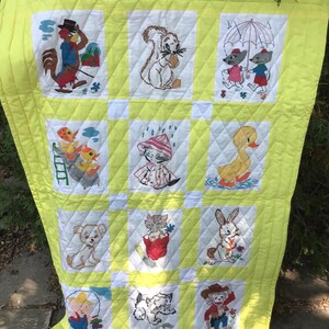 May include: A bright yellow quilted blanket featuring twelve white square panels, each embroidered with colourful illustrations of various animals including a rooster, squirrel, mice with an umbrella, ducklings, a kitten, a puppy, and a monkey.
