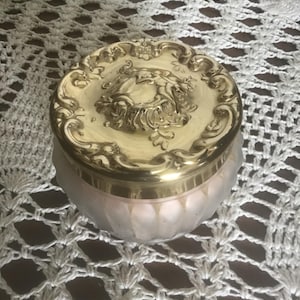 May include: A round glass jar with a frosted finish and vertical fluted detailing, topped with a polished gold-tone metal lid. The lid features an ornate, raised relief design depicting cherubs or putti surrounded by intricate scrollwork.