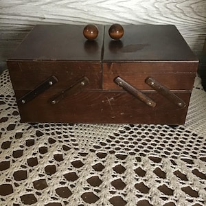 May include: A vintage wooden sewing box in a dark brown finish. The box features two hinged lids with round wooden knobs and pull-out drawers. It rests on a white crocheted doily.