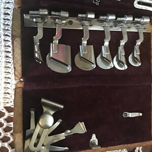 May include: A wooden case with a dark purple velvet interior containing a collection of silver-colored metal sewing machine attachments. The set includes various presser feet and hemmer guides arranged in rows, with some pieces secured on a metal bar.