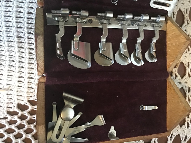 May include: A wooden case with a dark purple velvet interior containing a collection of silver-colored metal sewing machine attachments. The set includes various presser feet and hemmer guides arranged in rows, with some pieces secured on a metal bar.