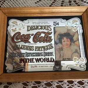 Puede incluir: Anuncio enmarcado de Coca-Cola con el texto "Delicious Coca-Cola" y "Relieves Fatigue". El letrero vintage incluye una imagen de una mujer y el texto "The Most Refreshing Drink in the World."