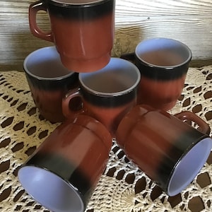 Op de afbeelding: Een verzameling van zes vintage koffiemokken. De mokken hebben een gradiëntontwerp, dat overgaat van een roodbruine basis naar een zwarte band aan de bovenkant, met een lichtpaars interieur. Elke mok heeft een handvat.