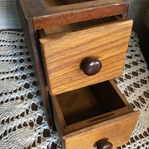 Op de afbeelding: Een vintage houten opbergdoos met twee lades, elk met een donkerbruine knop. De doos heeft een donkerbruine buitenkant en een lichtere houten binnenkant. De laden zijn gedeeltelijk open.