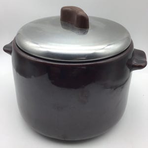 May include: A dark brown ceramic pot with a shiny silver lid. The lid has a small, brown handle. The pot has two side handles and a rounded shape. This kitchenware item is suitable for cooking and serving.