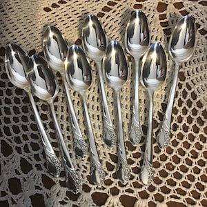Puede incluir: Una colección de nueve cucharas plateadas dispuestas sobre un mantel individual de encaje blanco. Las cucharas tienen una superficie reflectante y detalles florales ornamentados en los mangos. Las cucharas están dispuestas en forma de abanico.
