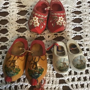 Peut inclure: Trois paires de sabots en bois miniatures. Une paire est rouge avec des motifs floraux blancs et le mot Holland. Une paire est en bois naturel avec une illustration de moulin et le mot Holland. Une paire est blanche avec un moulin.