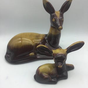 Op de afbeelding: Twee keramische hertenbeeldjes. Het grotere hert ligt, en het kleinere hert staat ervoor. De herten zijn geglazuurd in bruin- en beigetinten, met lange oren en een strak ontwerp. De beeldjes zijn waarschijnlijk decoratief.