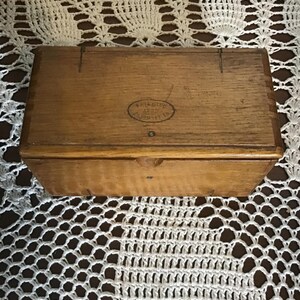 May include: A rectangular wooden box with a hinged lid, featuring a natural light brown wood grain finish. An oval stamp on the top lid displays the text PATENTED 1889 FEBRUARY 19. The box has visible joinery details along the edges and a small notch.