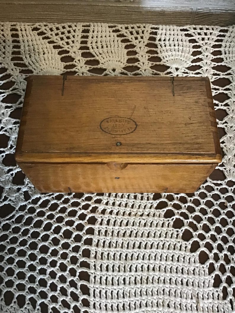 May include: A rectangular wooden box with a hinged lid, featuring a natural light brown wood grain finish. An oval stamp on the top lid displays the text PATENTED 1889 FEBRUARY 19. The box has visible joinery details along the edges and a small notch.