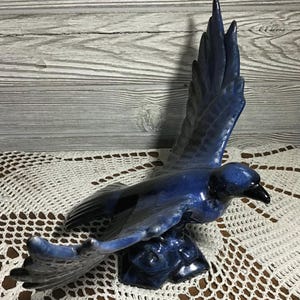May include: A ceramic bird sculpture in shades of blue and black, depicted mid-flight with outstretched wings. The sculpture is mounted on a textured base and displayed on a white lace doily. The bird's wings are spread wide.
