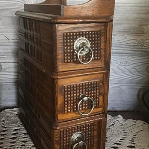 Puede incluir: Un cofre de almacenamiento vertical de madera con tres cajones apilados que presentan un patrón de rejilla texturizado en los paneles frontales. Cada cajón cuenta con un tirador de anillo metálico sujeto a una placa decorativa plateada.