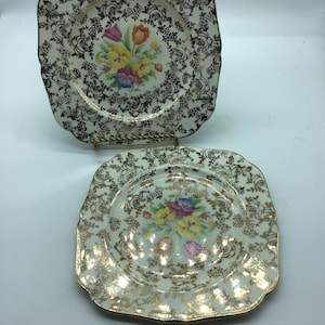 May include: Two square ceramic plates with a white background and gold trim. The plates feature a floral design with colourful flowers and leaves. The plates are ideal for serving desserts or snacks.