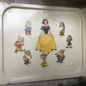 以下が含まれることがあります： 白の金属製のトレイに、白雪姫と7人の小人たちの絵が描かれています。トレイには、黄色のドレスを着た白雪姫と、それぞれ特徴的な色の7人の小人のカラフルなイラストが描かれています。トレイの上部には「Snow White」という文字が印刷されています。