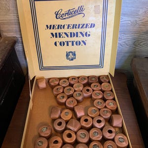 Könnte beinhalten: Ein alter Pappkarton mit zahlreichen kleinen Spulen aus braunem, mercerisiertem Stopfgarn. Der Deckel ist mit blauem Text bedruckt, der Corticelli, Mercerized Mending Cotton, Made in Canada lautet, und zeigt eine kleine Katzenabbildung.