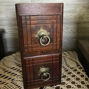 Könnte beinhalten: Ein dunkelbrauner Holzschrank mit zwei Schubladen. Jede Schublade hat einen dekorativen Messinggriff mit Ringgriff. Der Schrank hat eine rechteckige Form und ist aus Holz mit einem detaillierten Design.