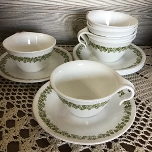 May include: White teacups and saucers with a green floral pattern. The teacups have a curved handle and are stacked in a group. The saucers and cups are arranged on a crocheted surface.