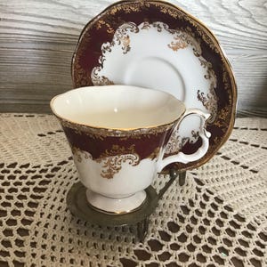Peut inclure: Une tasse et une soucoupe vintage. La tasse et la soucoupe ont une base blanche avec un motif floral bordeaux et or. La tasse est sur un petit support en métal, posé sur un napperon en crochet crème.