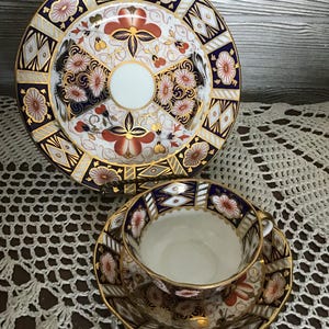 Peut inclure: Un service à thé en porcelaine vintage comprenant une tasse, une soucoupe et une assiette de service. L'ensemble présente une base blanche avec des motifs floraux et géométriques orange, bleu marine et or. Les bords sont dorés.