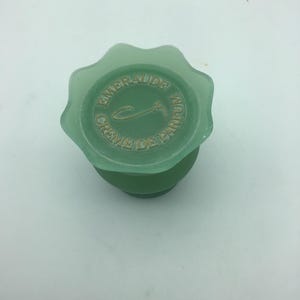 Może przedstawiać: Zielony szklany korek do butelki perfum z napisem "EMERAUDE CREME DE PARFUM" w złotych literach. Korek ma falisty brzeg i okrągłą górę.