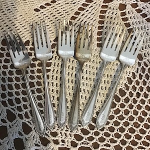 Peut inclure: Six fourchettes en argent vintage disposées sur un napperon en dentelle blanche. Les fourchettes présentent un design classique avec des dents et de longs manches. Chaque manche est orné d'un motif décoratif et d'un monogramme. Les fourchettes sont en argent brillant et terni.