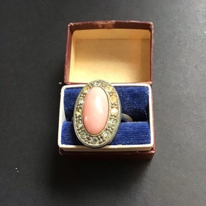 May include: A pink oval ring with a silver setting and clear rhinestones. The ring is in a blue velvet ring box.