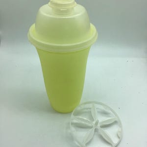 Puede incluir: Una coctelera de plástico amarillo pálido con tapa a juego y un colador transparente en forma de estrella. La coctelera tiene una base redondeada y una parte superior ligeramente ensanchada. La tapa es en forma de cúpula con un pequeño pico.