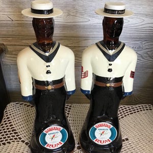Könnte beinhalten: Zwei Vintage-Rumflaschen aus Barbados in Form von Matrosen. Die Flaschen sind braun mit weiß und schwarz bemalten Uniformen und weißen Hüten mit der Aufschrift "Harbour Police". Jede Flasche hat ein Etikett mit der Aufschrift "Barbados Doorly's Rum".