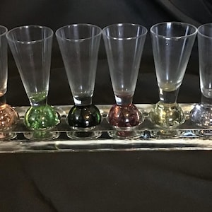Puede incluir: Un juego de seis vasos de chupito de vidrio transparente con bases redondeadas de colores en tonos naranja, verde, negro, rojo, amarillo y azul. Los vasos están dispuestos sobre una bandeja rectangular transparente.