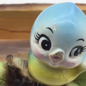 May include: A blue and yellow ceramic bird figurine with large eyes and a pink beak. The bird is perched on a brown branch with green leaves.