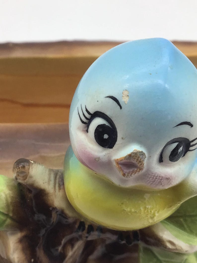 May include: A blue and yellow ceramic bird figurine with large eyes and a pink beak. The bird is perched on a brown branch with green leaves.