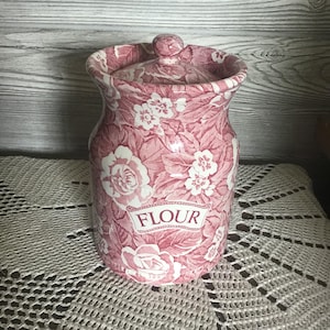 May include: A pink and white floral ceramic flour canister with a lid. The canister has a curved shape and the word "FLOUR" is printed on the front. The floral pattern includes roses and other flowers.