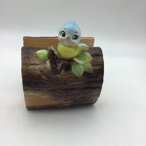 May include: A ceramic napkin holder shaped like a tree log with a blue bird perched on a branch. The bird has yellow feathers and large, black eyes.
