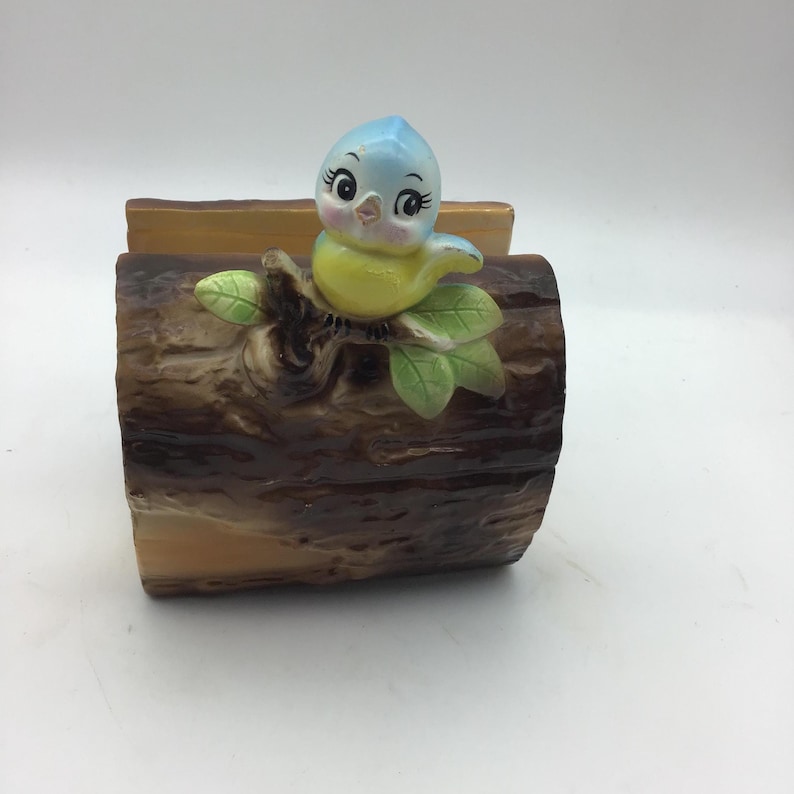 May include: A ceramic napkin holder shaped like a tree log with a blue bird perched on a branch. The bird has yellow feathers and large, black eyes.