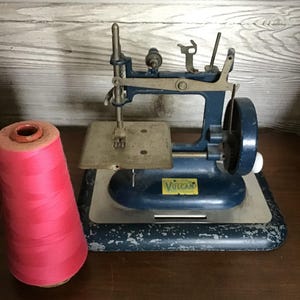 May include: A vintage blue Vulcan sewing machine with a metal base and a yellow brand label, positioned next to a large cone of bright pink thread. The machine features a hand crank wheel and a metallic needle plate with visible signs of wear.