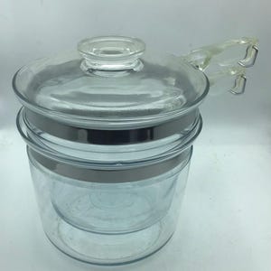 May include: A clear glass double boiler pot featuring a matching glass lid with a central knob handle. The set includes a top insert and a bottom vessel, both encircled by a polished silver-tone metal band. A clear glass handle extends from the side.