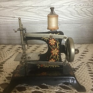 May include: A vintage black metal sewing machine featuring hand-painted orange and yellow floral motifs on the frame. The machine includes a silver-toned hand crank wheel, a needle bar assembly, and a wooden spool of light beige thread on top.
