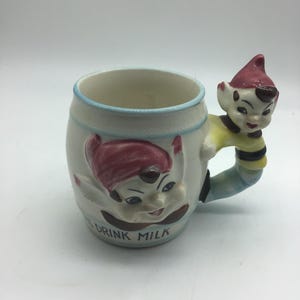 Puede incluir: Una taza de cerámica blanca con una franja azul y blanca alrededor de la parte superior. La taza tiene un asa con forma de elfo con un gorro rojo, una camisa amarilla y rayas negras. La cara del elfo está pintada en la taza, y las palabras "Drink Milk" están escritas en la parte delantera.
