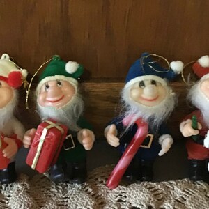 May include: Four vintage Christmas tree decorations shaped like elves. Each elf is wearing a different coloured hat: red, green, blue, and white. The elves are holding different Christmas-themed items, such as a candy cane, a gift box, and a scroll.