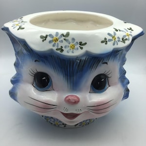 May include: A ceramic cat-shaped planter with blue and white detailing. The planter has large blue eyes, pink cheeks, and a smiling mouth. The rim is decorated with small blue and yellow flowers. A decorative item for the home.