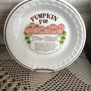以下が含まれることがあります： 茶色の縁と茶色の文字で「PUMPKIN Pie」と書かれた白いセラミックパイ皿。 皿にはピンクのかぼちゃのグラフィックとレシピが描かれています。 皿は装飾的なスタンドにあります。
